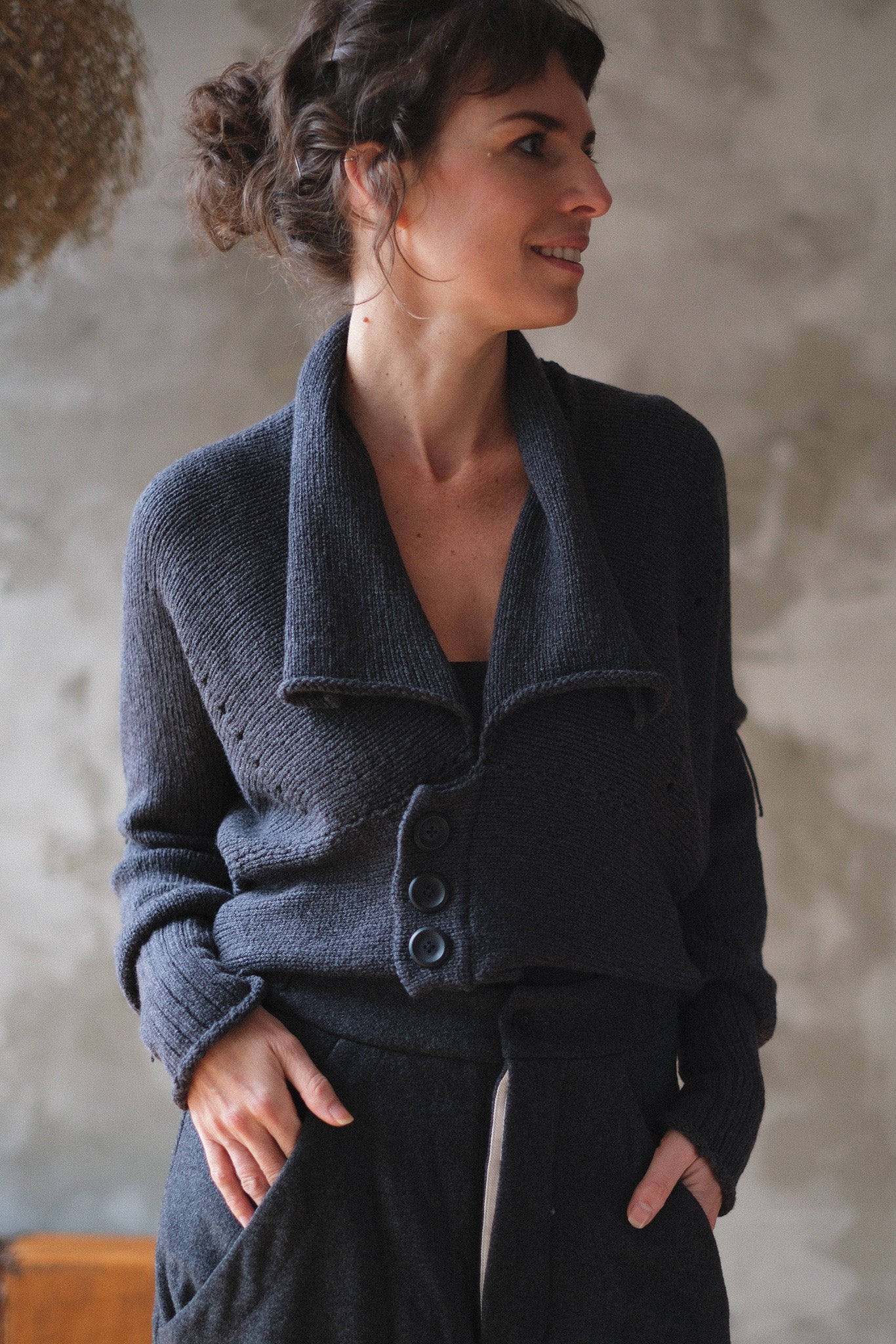 A woman wearing a charcoal gray knit cardigan with a wide collar and three front buttons, paired with matching dark trousers, standing against a textured plaster wall.