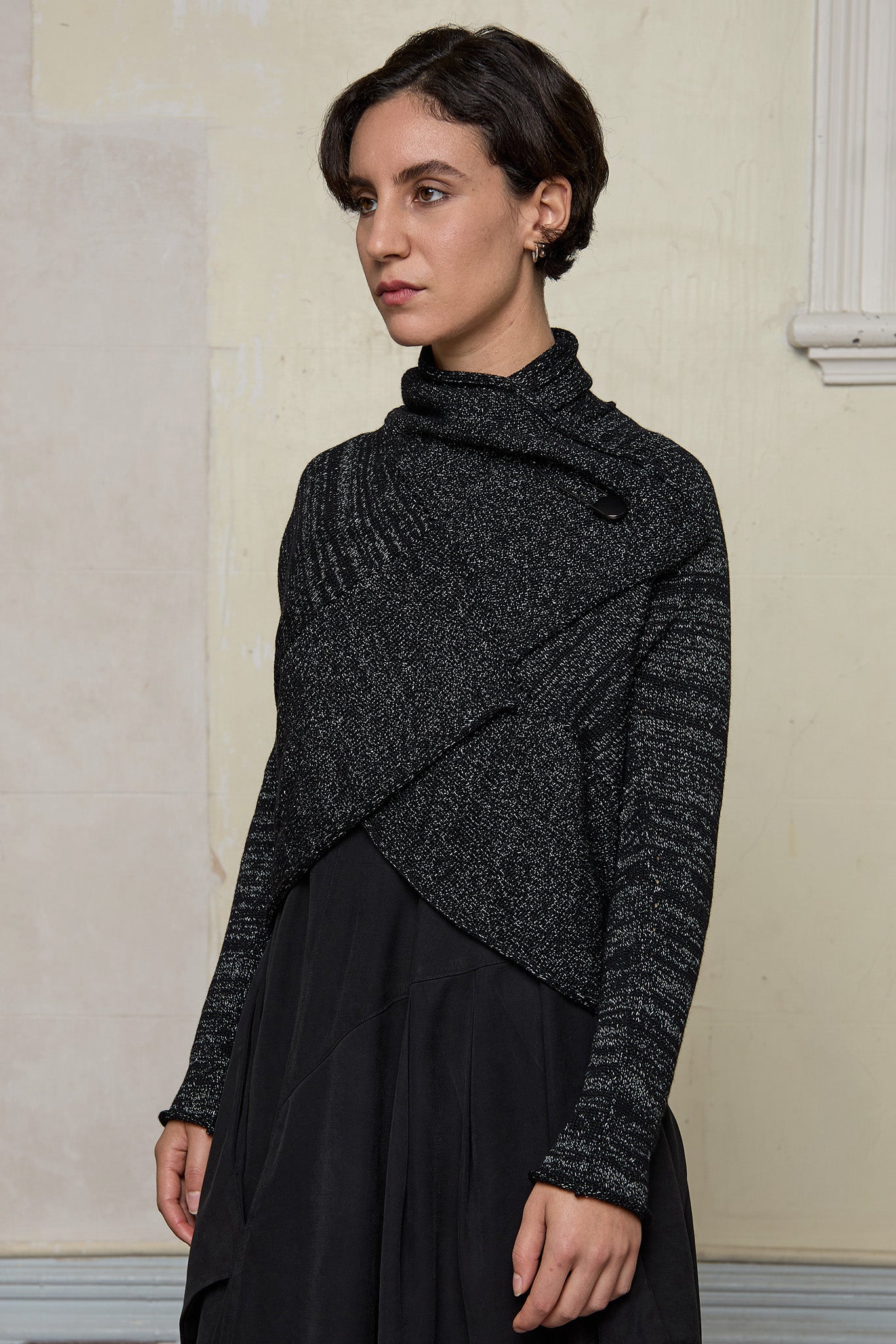 Model wears a black with white flecks shawl collared cardigan upside down, and fastened high at the neck using a safety pin.