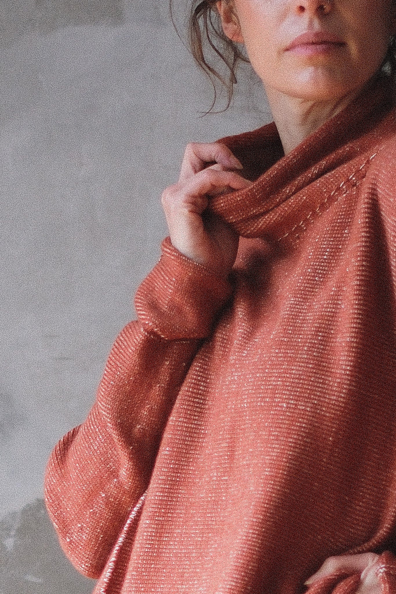 A close-up of a person wearing a textured, erracotta knit sweater with a high draped neckline, set against a neutral gray concrete wall.