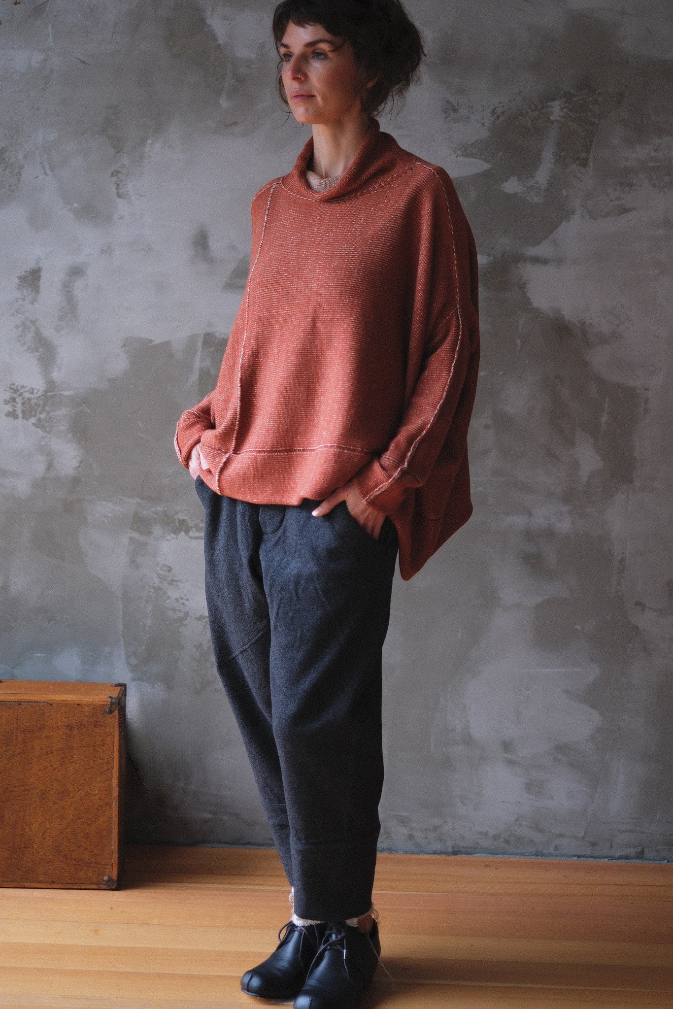 A woman wearing a loose, terracotta cowl-neck knit sweater, dark charcoal pants, and black boots, standing against a textured gray concrete wall.