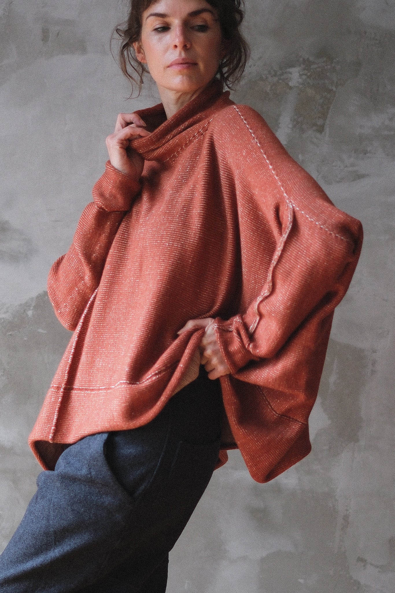 A woman wearing a loose, oversized knit poncho sweater in terracotta, paired with dark blue jeans, posed against a textured gray concrete wall.