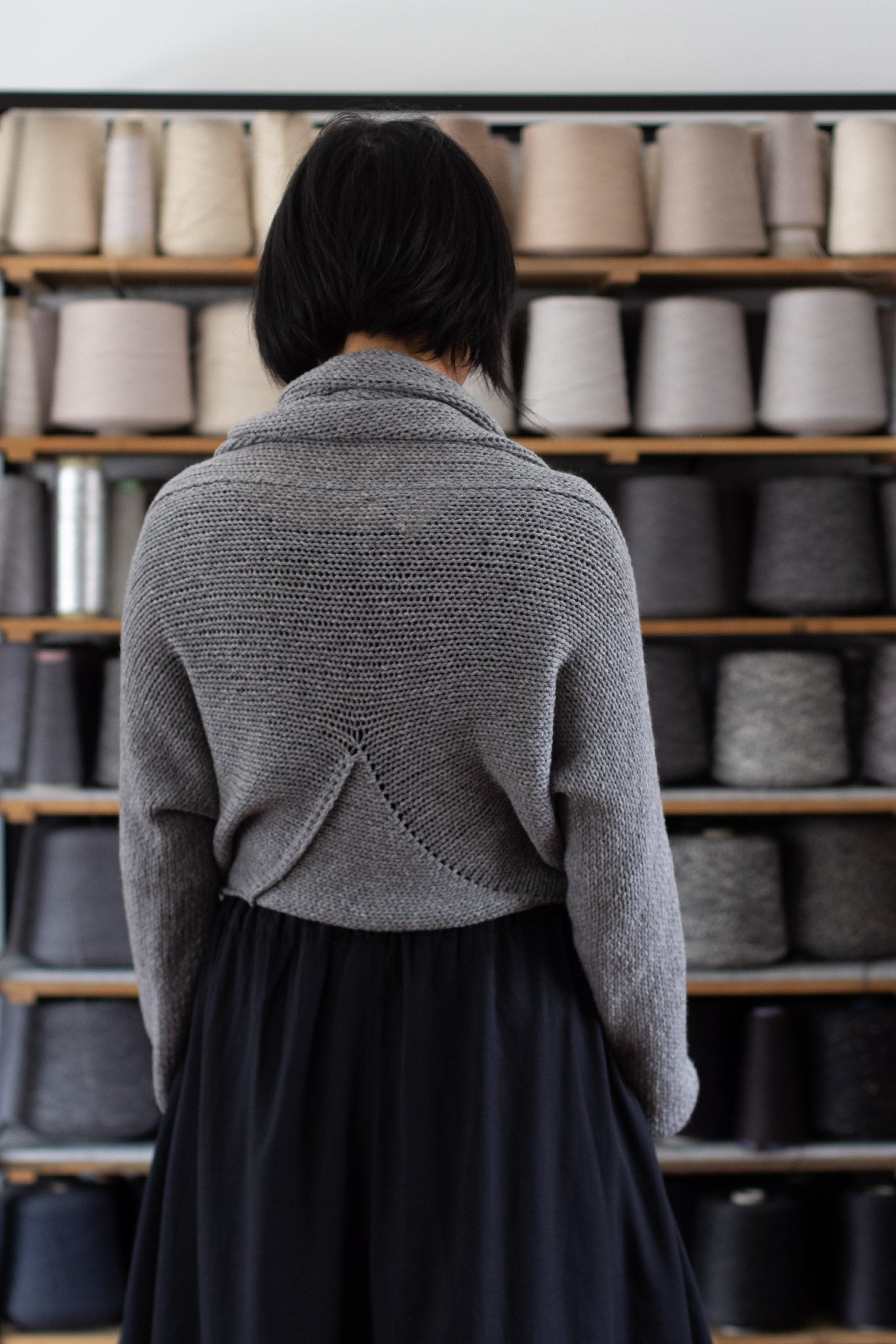 Model is standing in front of a shelf of yarns, showing the back view of a chunky knit merino wool jumper in soft grey, designed and made in Melbourne by Wendy Voon