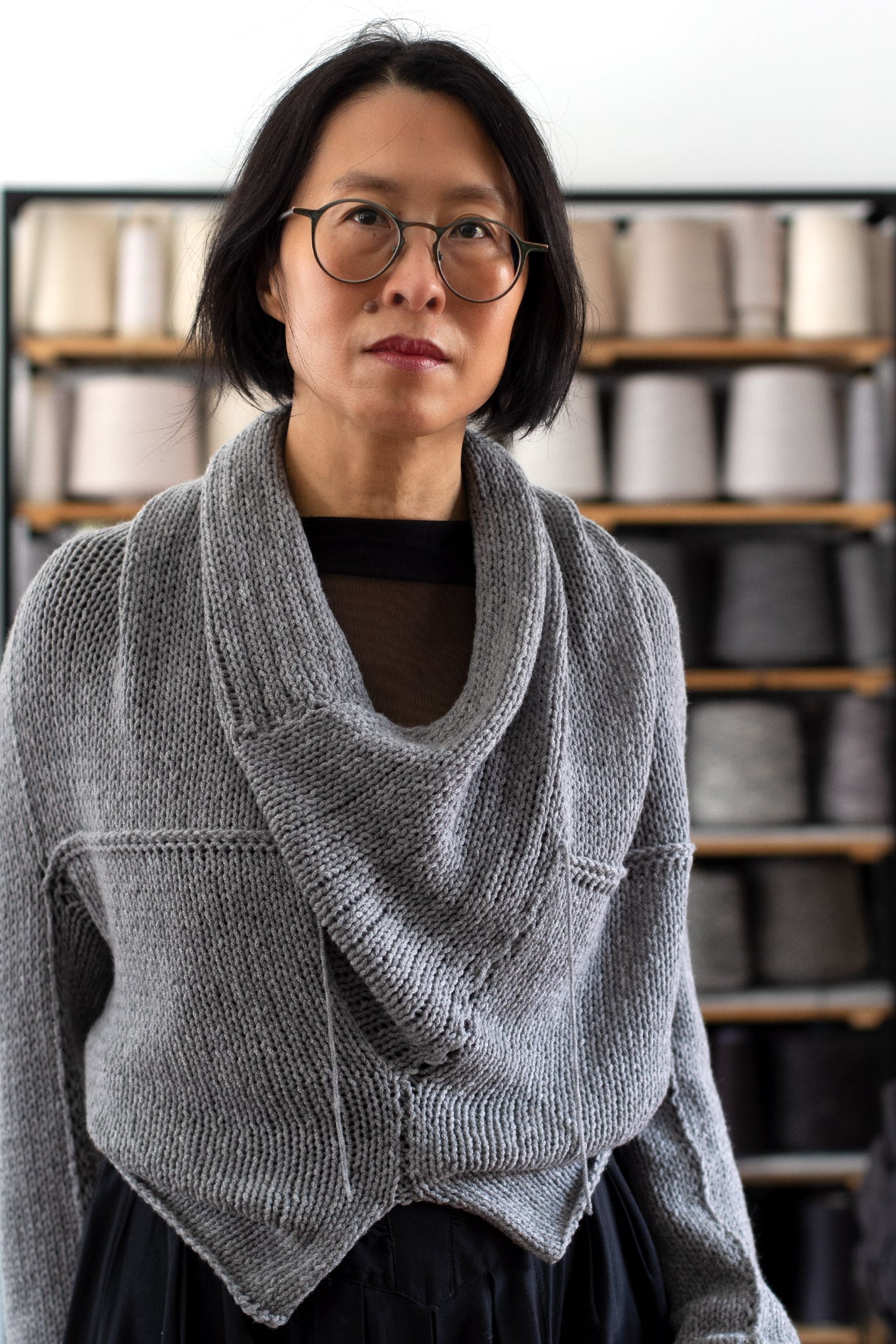 Model is standing in front of a shelf of yarns, wearing a chunky knit merino wool jumper in soft grey, designed and made in Melbourne by Wendy Voon