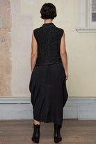 Back view of model wearing asymmetric self-curling vest in silver metallic on black merino wool vest, tied at the front.