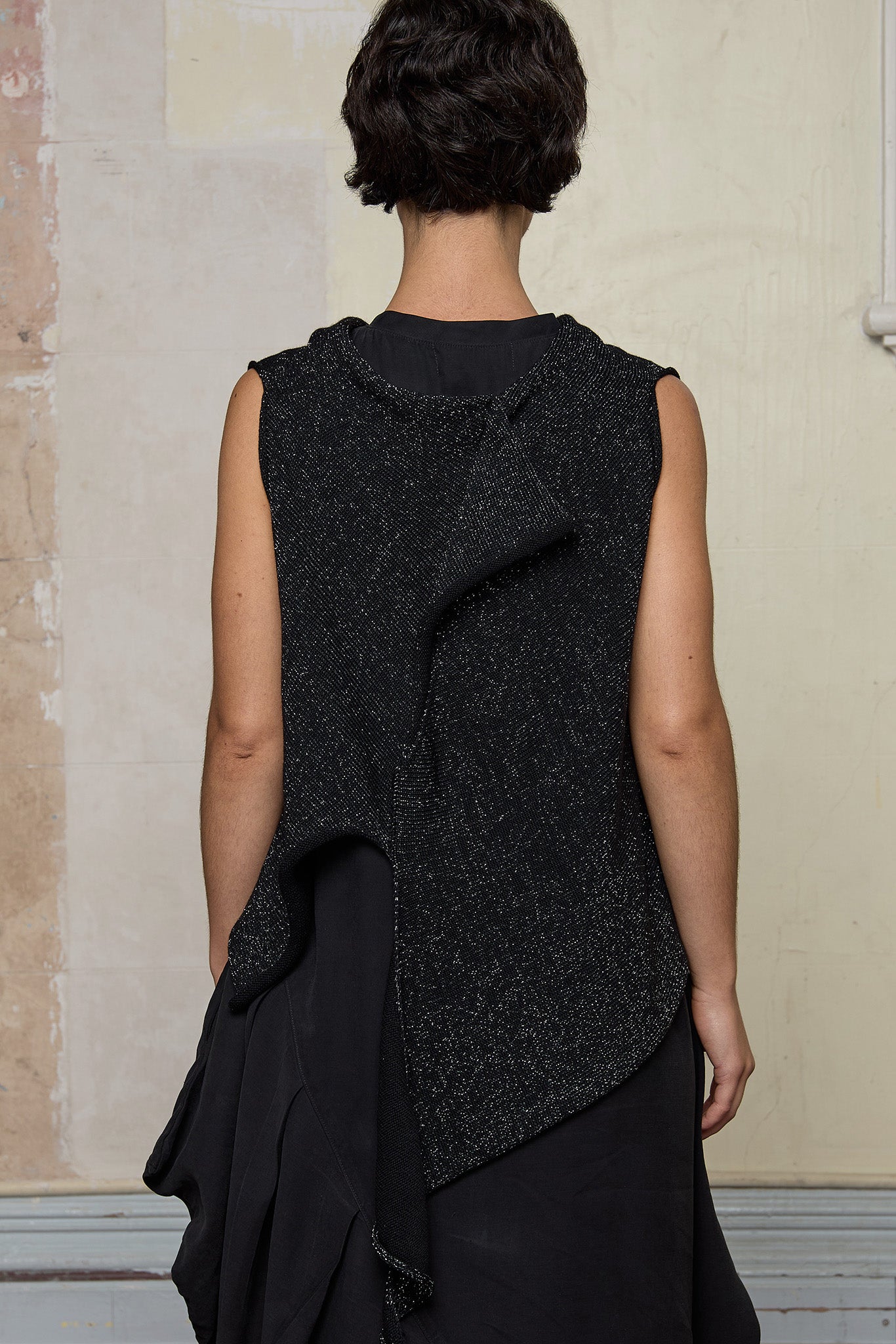 Back view of model wearing asymmetric self-curling vest back to front, in silver metallic on black merino wool vest.