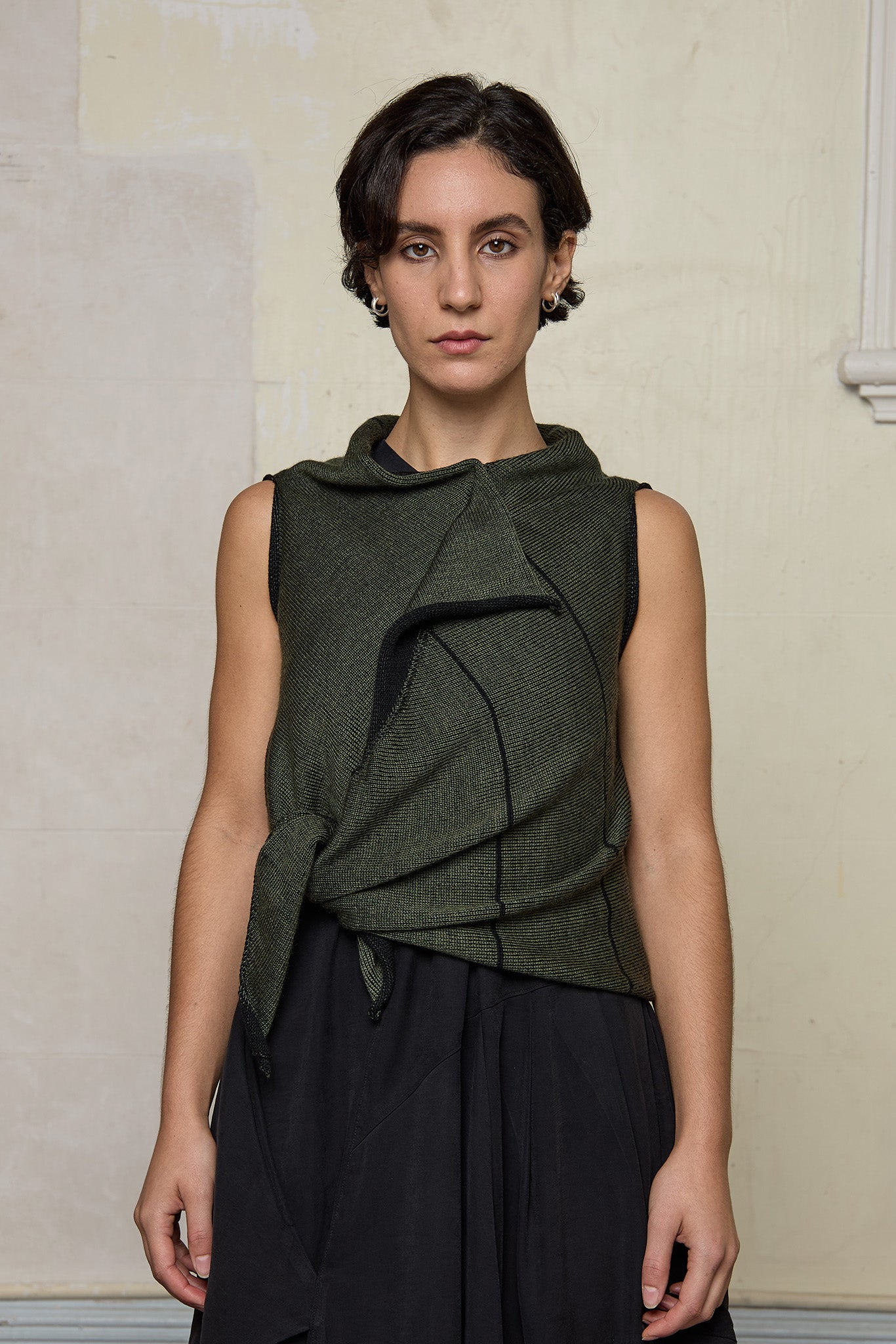 Model wears knitted asymmetric self-curling vest cinched at the waist, in army green on black colourway.