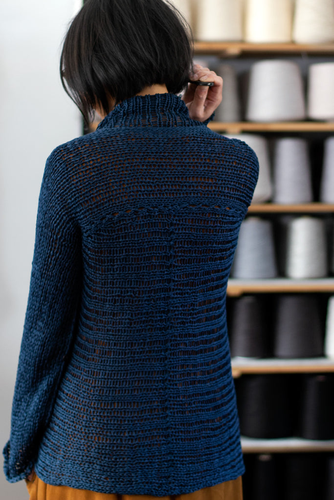 Back view of deep ocean coloured open knit cotton jumper, designed and knitted by Wendy Voon, with spools of yarn in the background.