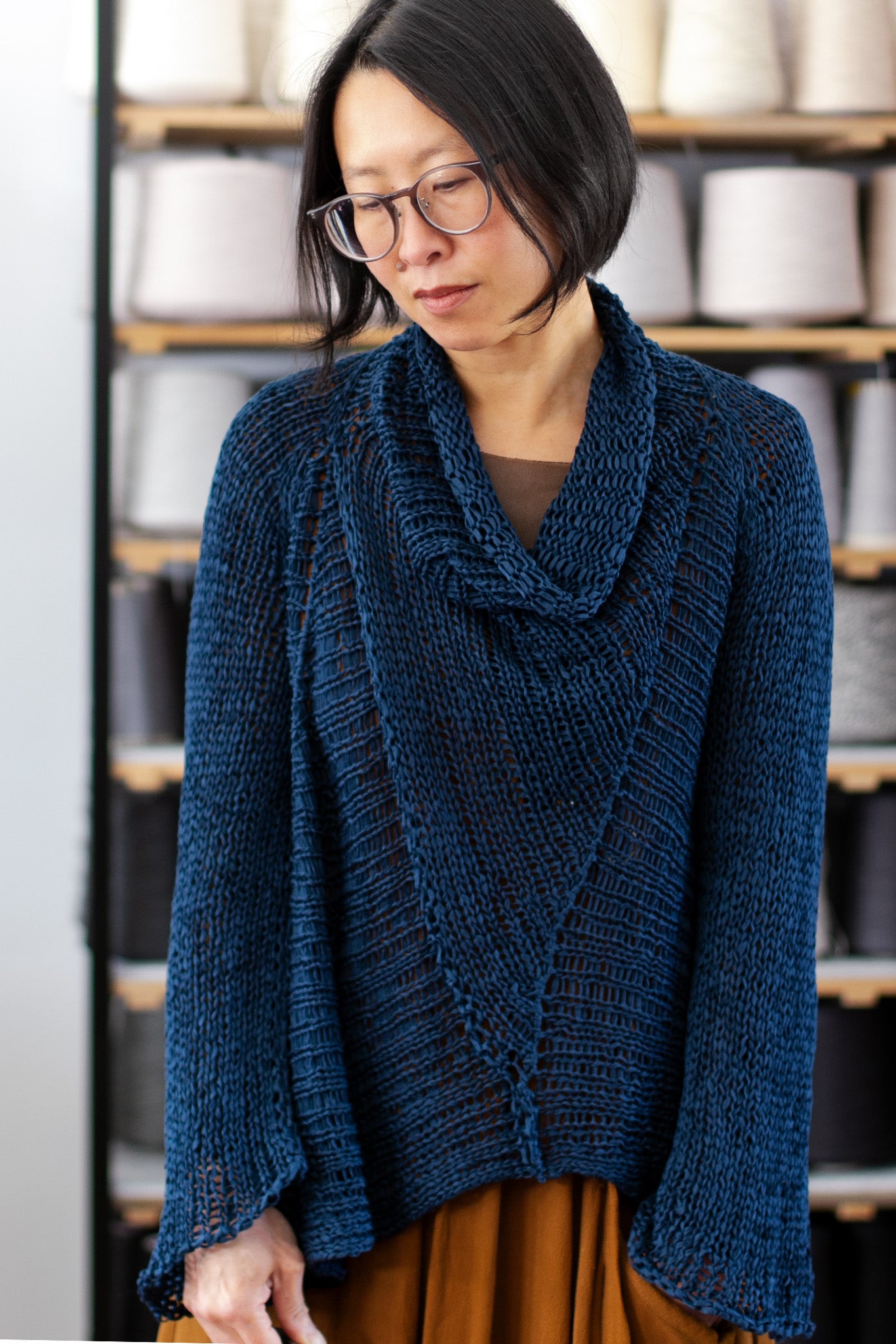 Front view of deep ocean coloured open knit cotton jumper with funnel neck, designed and knitted by Wendy Voon, with spools of yarn in the background.
