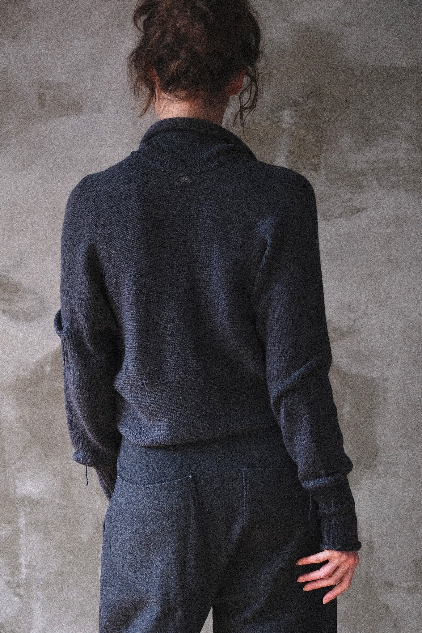 A person shown from the back wearing a charcoal gray knit sweater and matching dark trousers, standing against a textured gray wall.