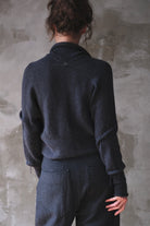 A person shown from the back wearing a charcoal gray knit sweater and matching dark trousers, standing against a textured gray wall.