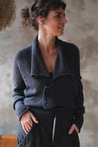 A woman wearing a charcoal gray knit cardigan with a wide collar and three front buttons, paired with matching dark trousers, standing against a textured plaster wall.