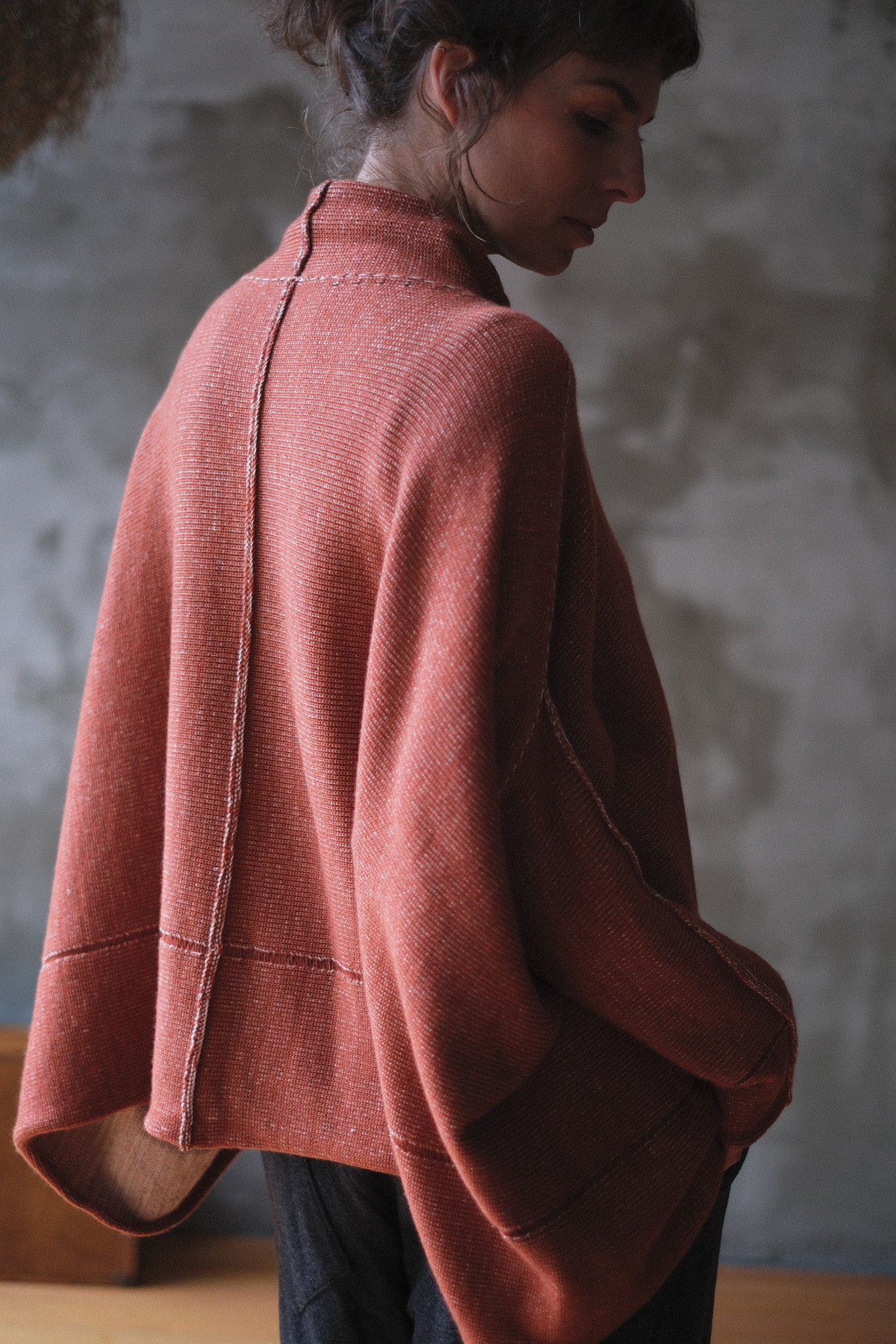 A woman shown from the back wearing a loose, draped knit jumper in a muted terracotta coloir, paired with dark pants, against a textured gray wall.
