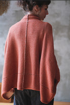 A woman shown from the back wearing a loose, draped knit jumper in a muted terracotta coloir, paired with dark pants, against a textured gray wall.