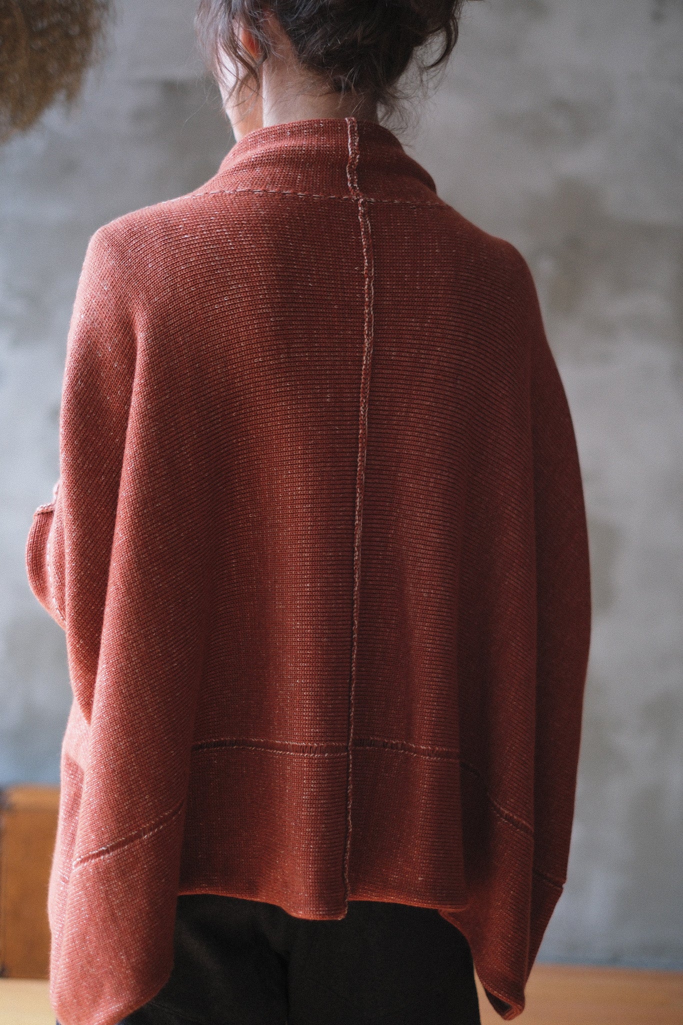 A woman shown from the back wearing a loose, draped knit jumper in a muted terracotta coloir, paired with dark pants, against a textured gray wall.