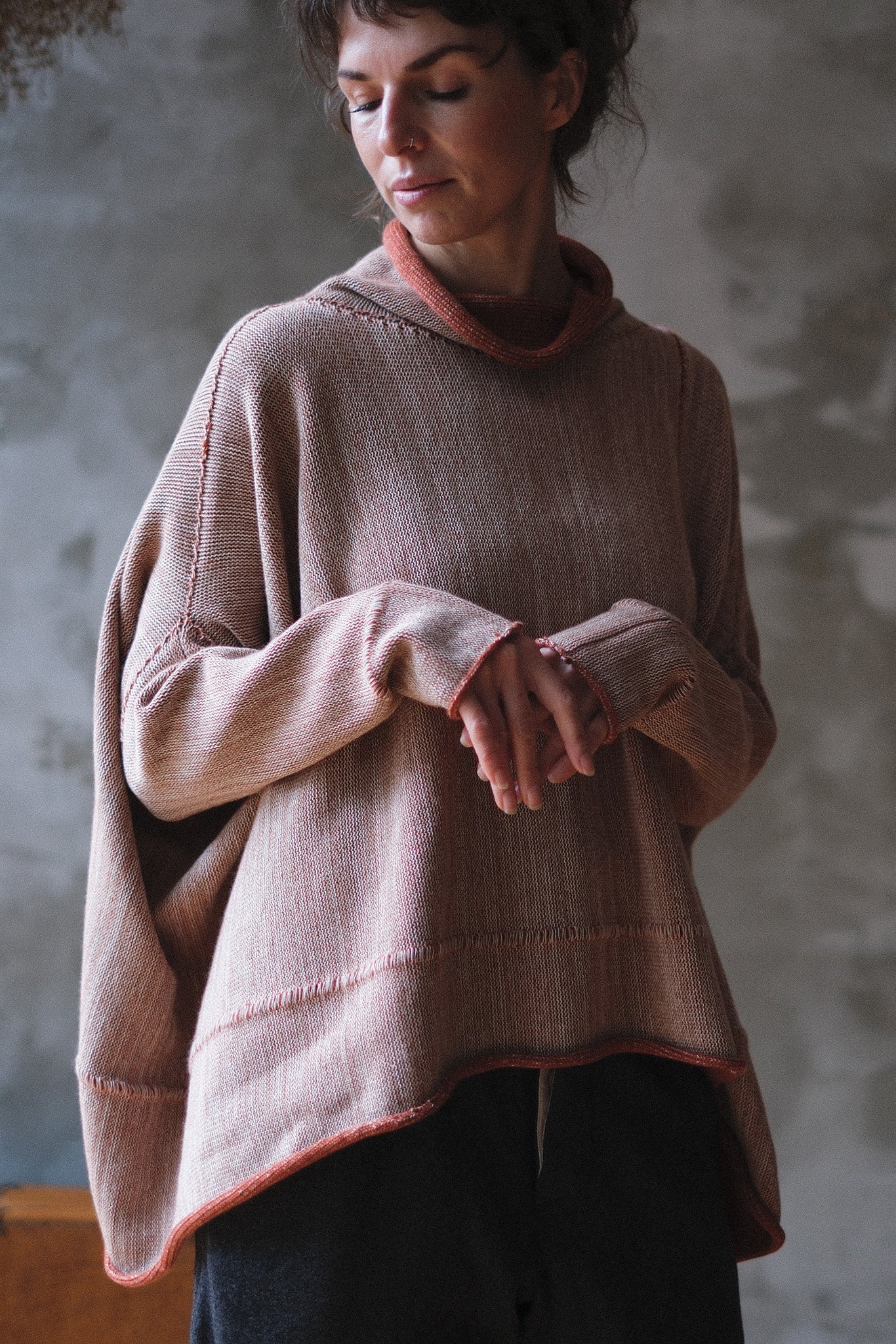 A woman wearing a loose, oversized knit poncho sweater in a fawn terracotta melange with a contrasting terracotta trim, standing against a textured gray concrete wall.