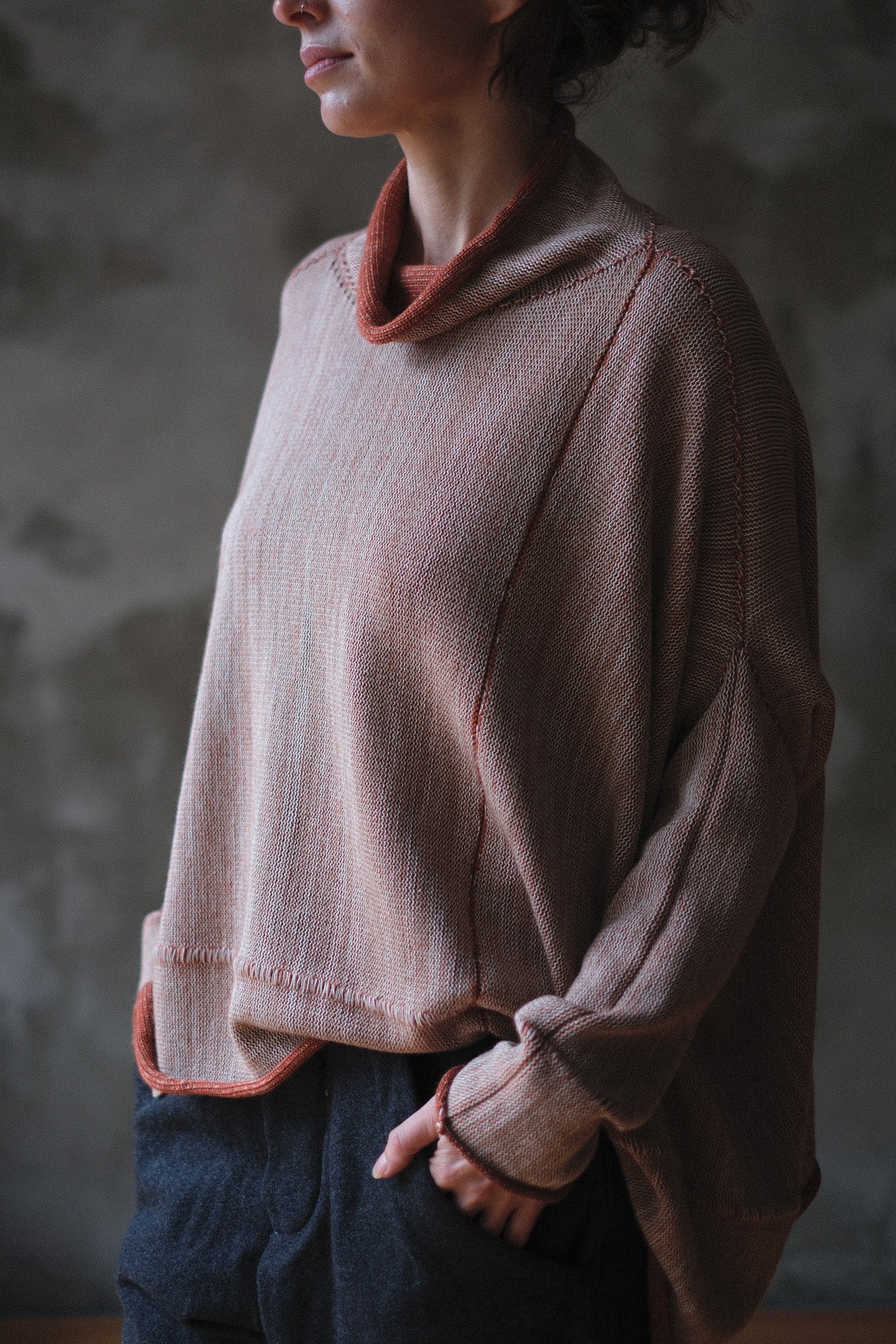 A person wearing a loose, oversized cowl-neck knit sweater in a fawn and terracotta melange colour with contrasting rust-orange trim, paired with dark pants, set against a textured gray wall.