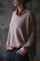 A person wearing a loose, oversized cowl-neck knit sweater in a fawn and terracotta melange colour with contrasting rust-orange trim, paired with dark pants, set against a textured gray wall.