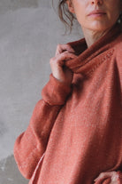 A close-up of a person wearing a textured, erracotta knit sweater with a high draped neckline, set against a neutral gray concrete wall.