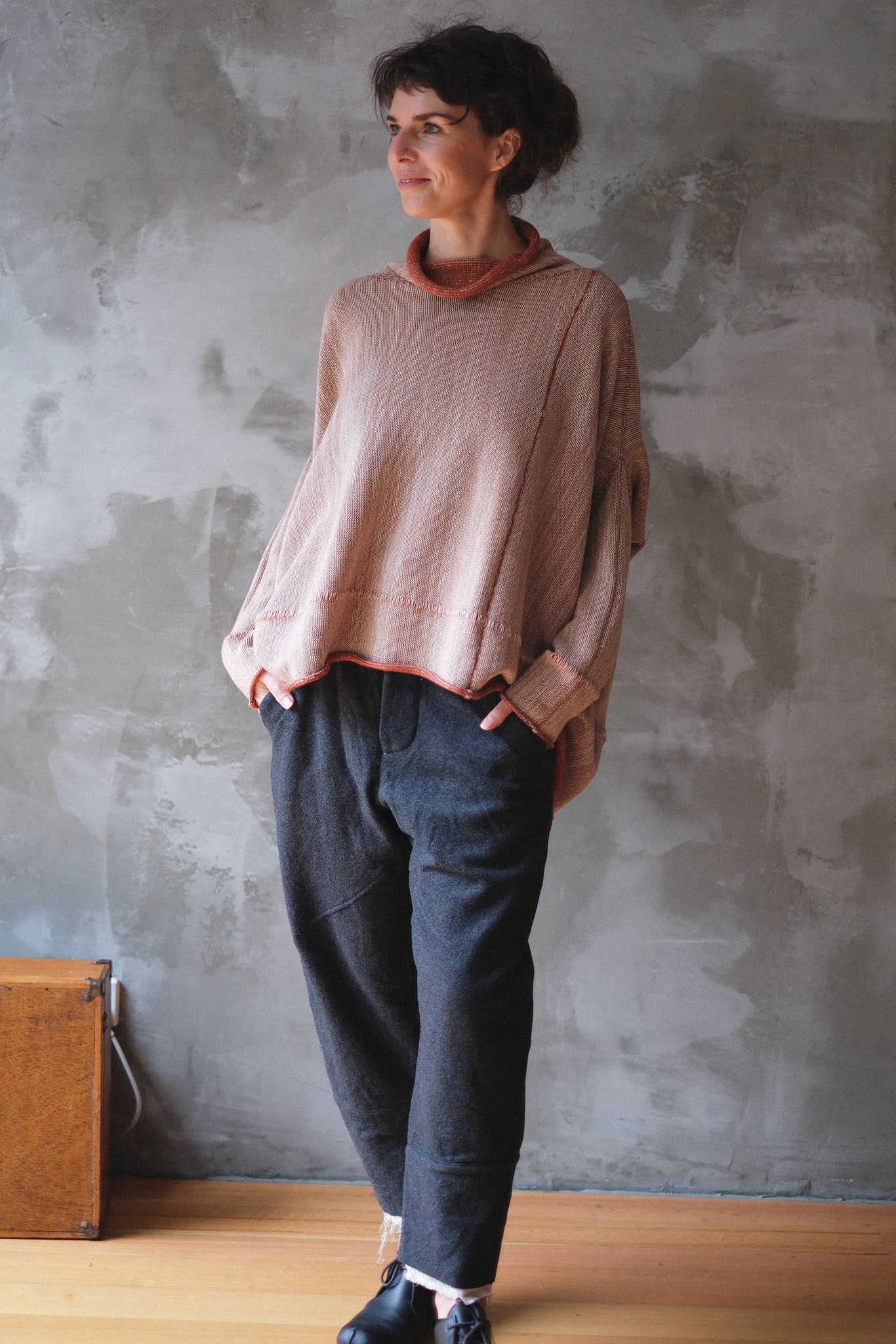 A woman wearing a loose, dusty fawn and terracotta melange knit sweater and dark charcoal gray trousers, standing against a textured gray concrete wall.
