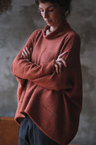 A woman wearing a loose, oversized cowl-neck knit sweater in a muted terracotta or rust color, paired with dark pants, standing with her arms crossed against a textured gray wall.