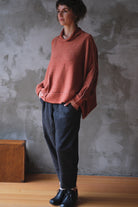 A woman wearing a loose, terracotta cowl-neck knit sweater, dark charcoal pants, and black boots, standing against a textured gray concrete wall.