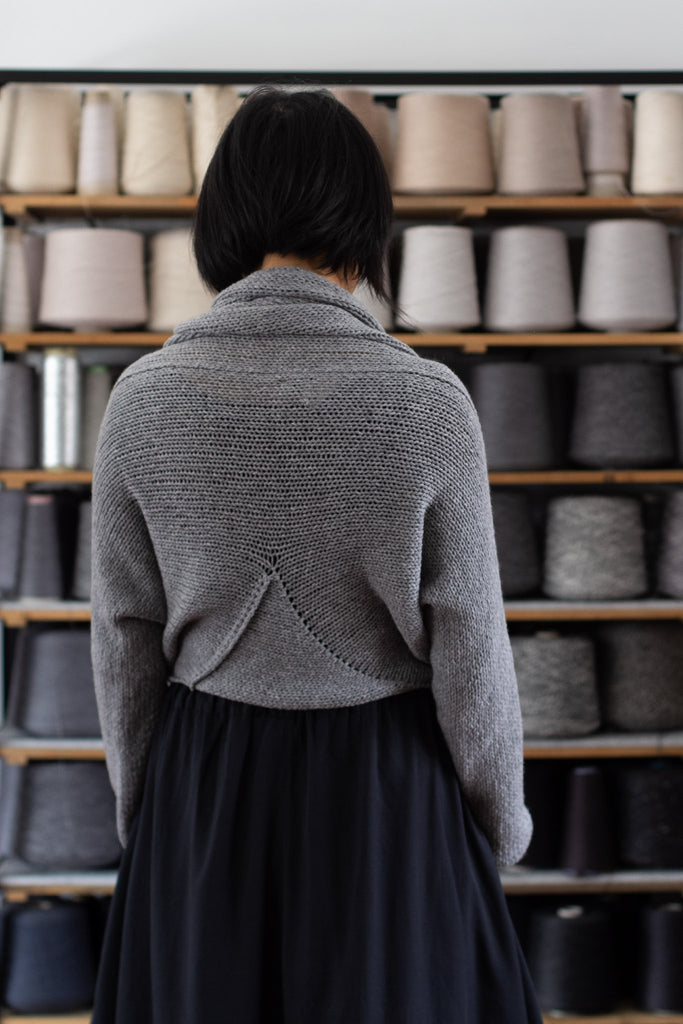 Model is standing in front of a shelf of yarns, showing the back view of a chunky knit merino wool jumper in soft grey, designed and made in Melbourne by Wendy Voon