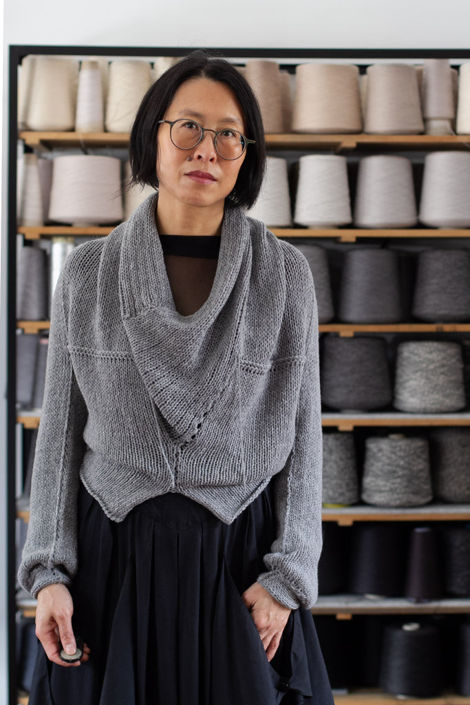 Model is standing in front of a shelf of yarns, wearing a chunky knit merino wool jumper in soft grey, designed and made in Melbourne by Wendy Voon