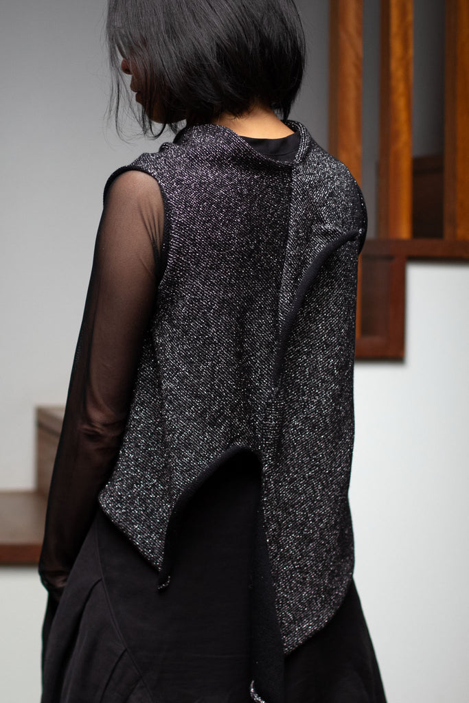 Back view of model wearing an asymmetric self curling vest back to front, in a silver metallic backed with black merino wool fabric.