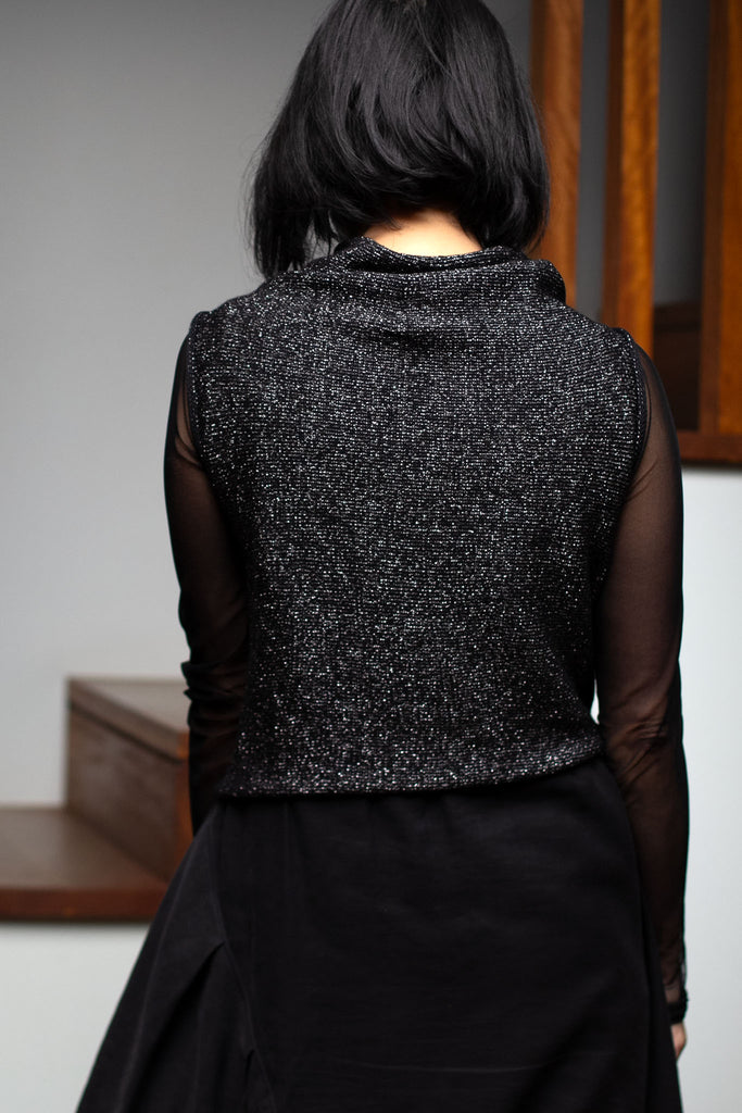 Back view of model wearing an asymmetric self curling vest, in a silver metallic backed with black merino wool fabric.