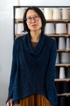 Front view of deep ocean coloured open knit cotton jumper with funnel neck, designed and knitted by Wendy Voon, with spools of yarn in the background.