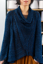 Front view of deep ocean coloured open knit cotton jumper with funnel neck, designed and knitted by Wendy Voon, with spools of yarn in the background.