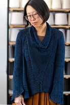Front view of deep ocean coloured open knit cotton jumper with funnel neck, designed and knitted by Wendy Voon, with spools of yarn in the background.