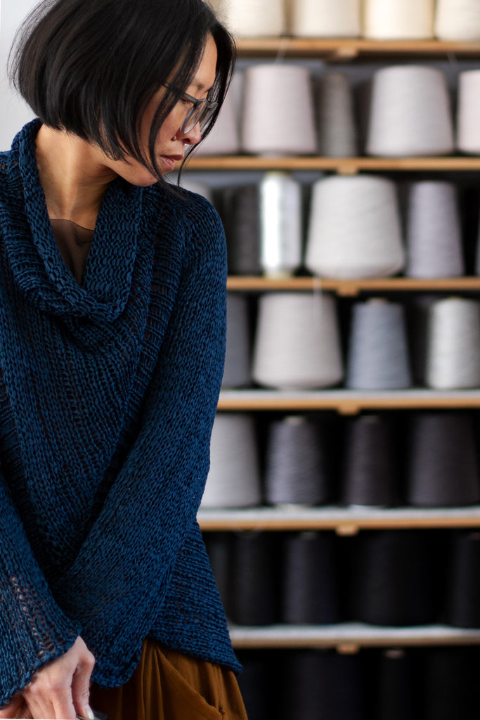 Partial view of deep ocean coloured open knit cotton jumper with funnel neck, designed and knitted by Wendy Voon, with spools of yarn in the background.